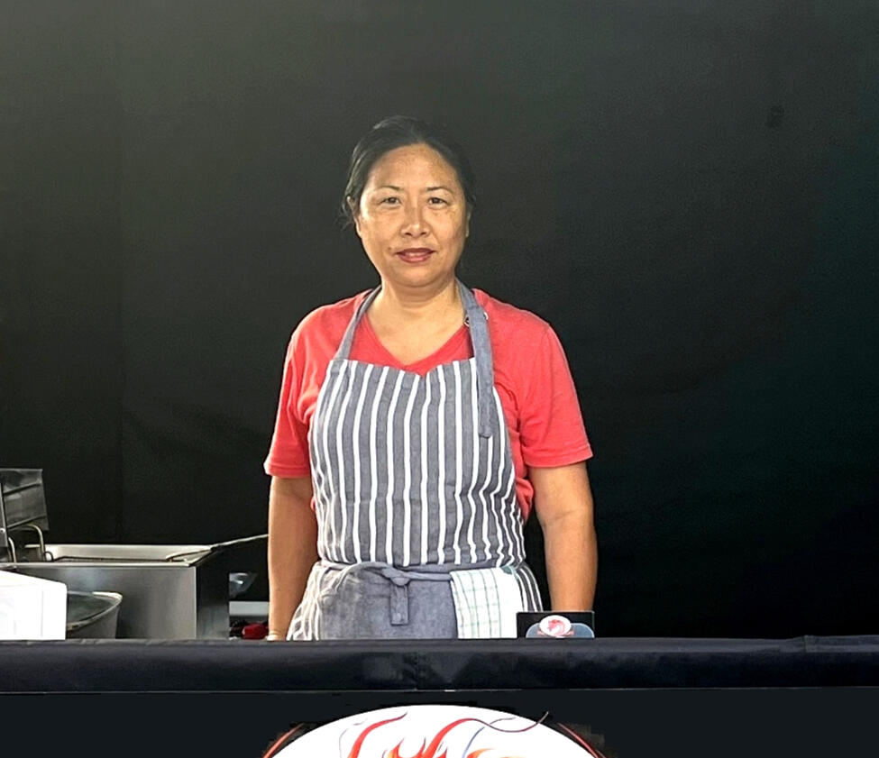 Chef Helen Portrait of Helen, the chef behind Oriental Savouries, standing at the market stall with a dark background and part of the dragon logo visible.